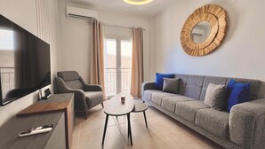 A modern living room with a flat-screen TV, a grey sofa with blue cushions, a grey armchair, a small round coffee table, and a wooden side table. - Fifth Sense in Athens by ΜΡS (Athina)