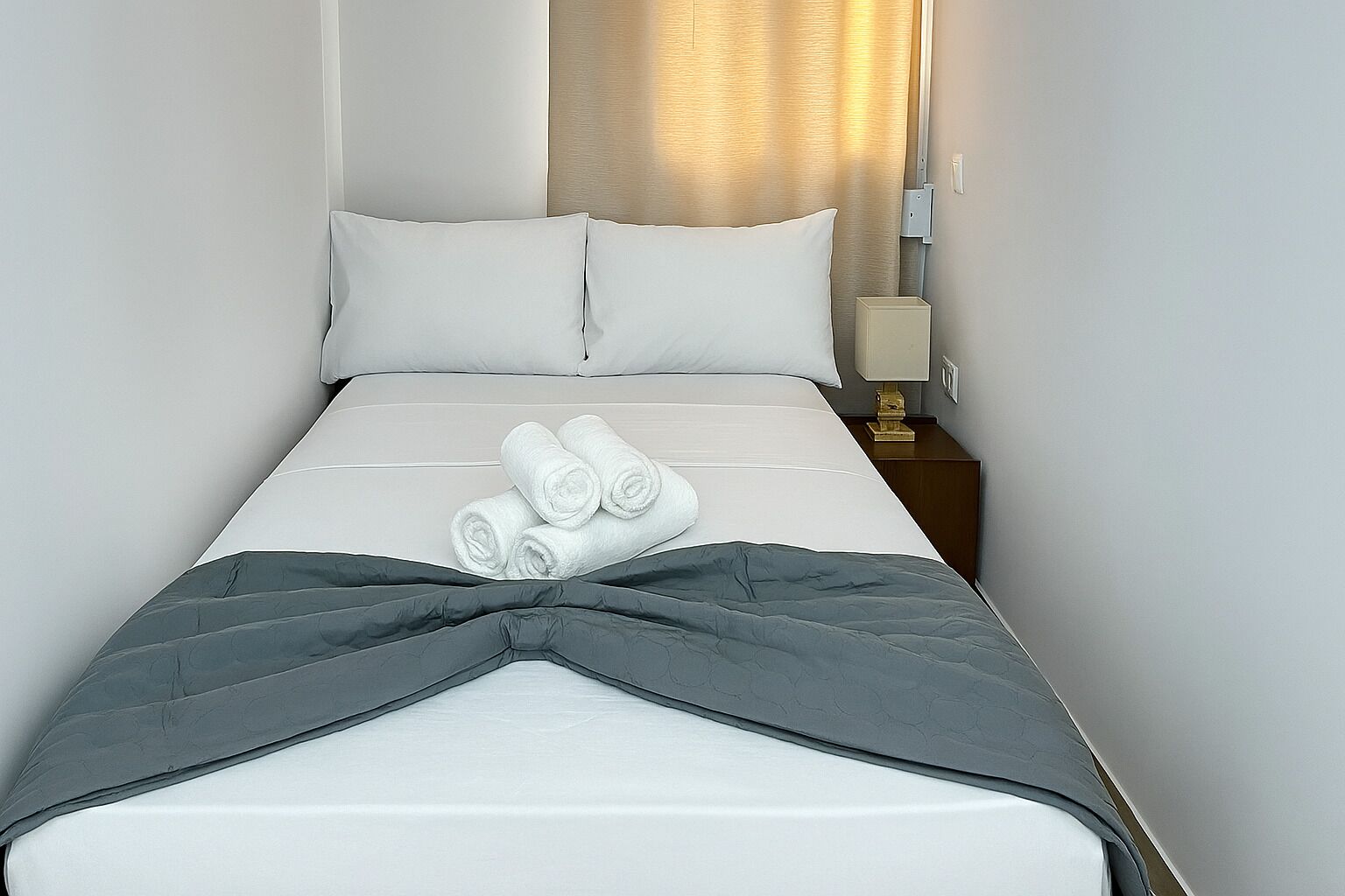 Un lit bien fait, avec du linge de lit blanc et une couverture grise, une lampe de chevet et un luminaire fixé au mur.