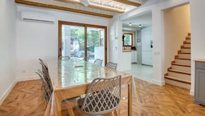 Un comedor con una gran mesa de madera, sillas y un ventilador de techo. Se observa una cocina al fondo.