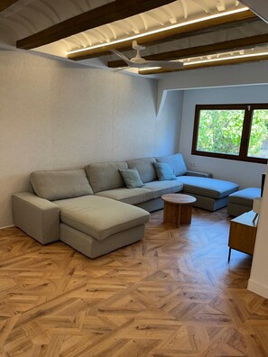 Un salon moderne avec un canapé d’angle, une table basse ronde en bois et une fenêtre donnant sur un paysage verdoyant.