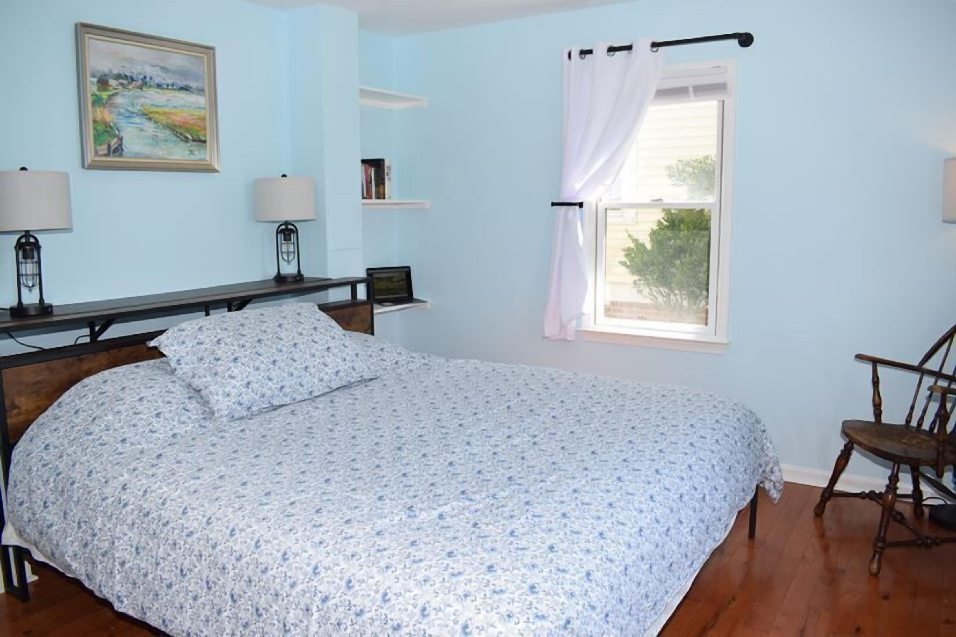 A bedroom with a bed, bedside lamps, a chair, a window with curtains, and a painting on the wall.