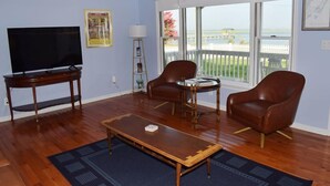 A living room with a TV, two leather chairs, a coffee table, and a large window with a view of the beach.