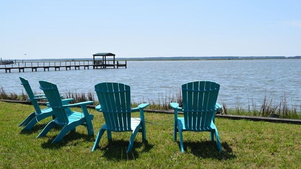 Three turquoise Adirondack chairs facing a body of water with a dock extending into it.