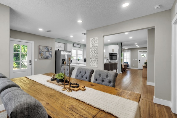 A spacious living area with a wooden dining table, grey upholstered sofas, and a kitchen in the background.