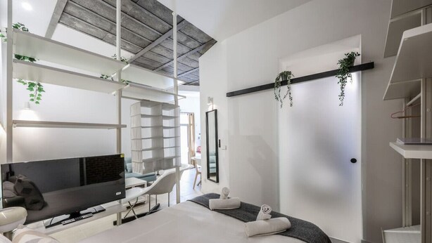 A modern hotel room with a bed, a desk, and a TV. There are plants on the wall and a staircase in the background.