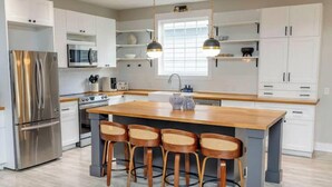 A modern kitchen with white cabinets, stainless steel appliances, a large island with bar stools, pendant lighting, and a window providing natural light. - Private Cinema + Cowboy Pool, Putt Putt, Arcade (Indianapolis)