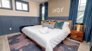 A neatly made bed with a white duvet, a patterned rug, a wooden nightstand, and a wall with the name 'INDY'. - Private Cinema + Cowboy Pool, Putt Putt, Arcade (Indianapolis)