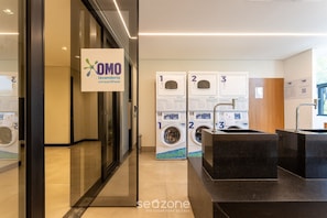A modern laundry facility with three washing machines labeled 1, 2, and 3, and a sign for OMO laundry detergent. - VNU - Apartamentos próximos de atrações em SP (São Paulo)