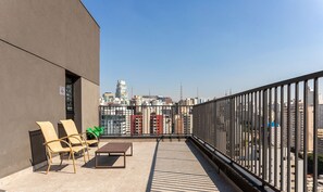 A rooftop terrace with a view of a city skyline, featuring a modern building, a railing, and outdoor furniture. - VNU - Apartamentos próximos de atrações em SP (São Paulo)