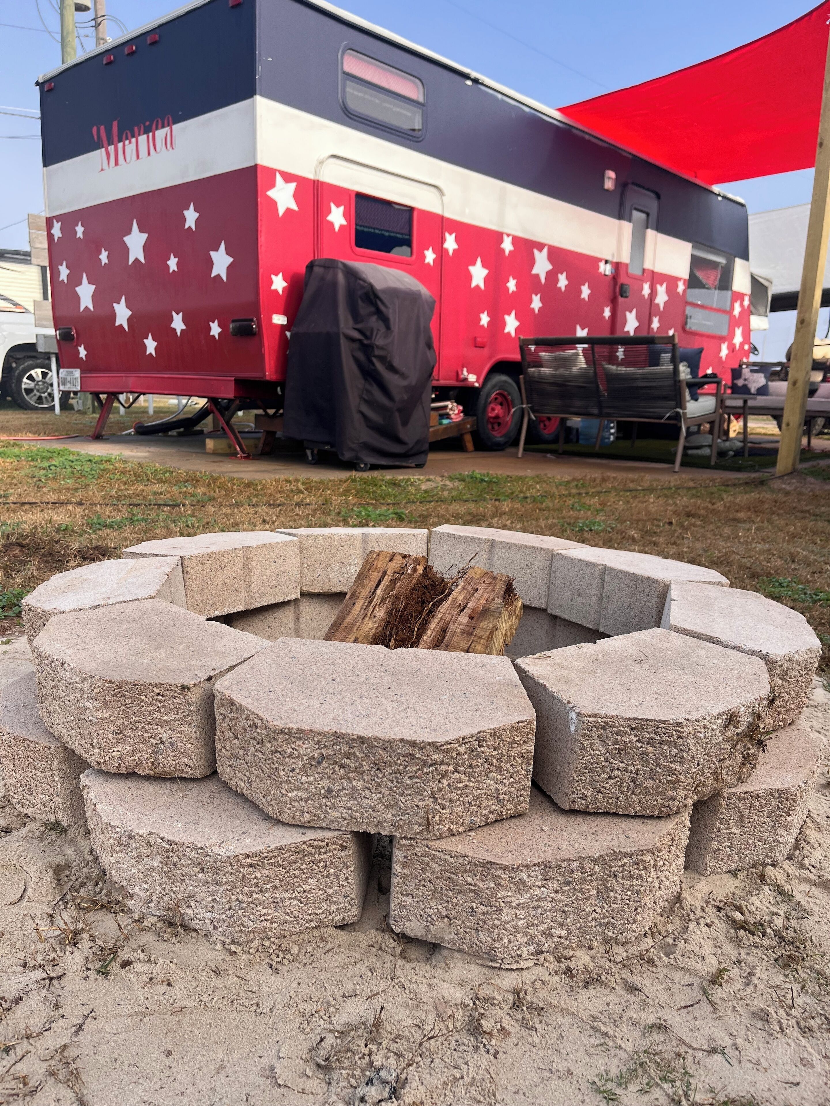 A fire pit made of hexagonal stones, a covered RV with 'America' written on it, and a red canopy.