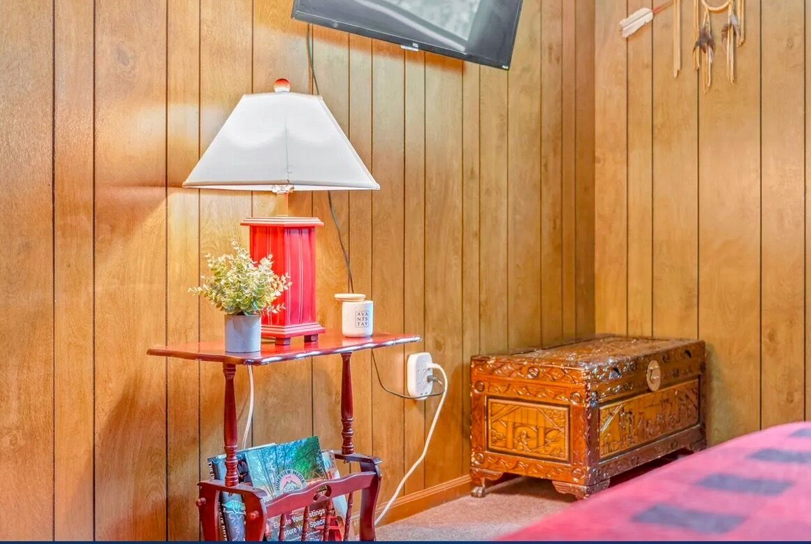Een kamer met houten lambrisering, een klein tafeltje, een lamp, een rode lampenvoet, een witte vaas met een plant, een klein houten kistje en een aan de muur bevestigde televisie.