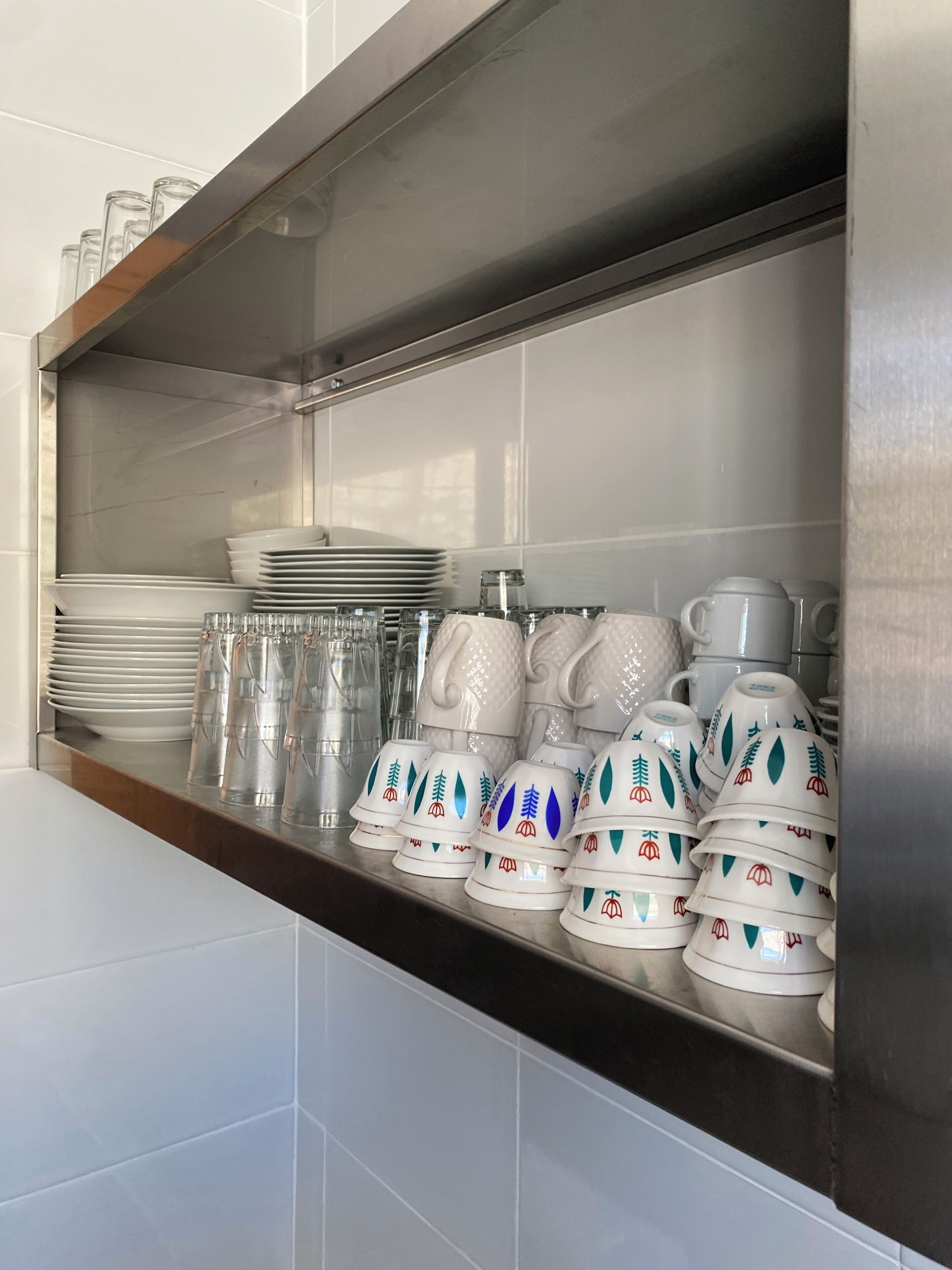 A kitchen area with stainless steel appliances, white tiled walls, and a shelf with stacked plates, glasses, and decorative cups.
