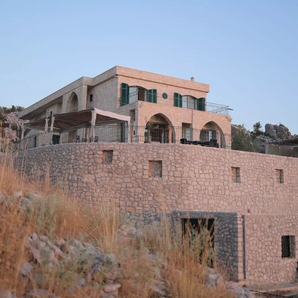 A multi-story stone building with a flat roof, arched windows, and a balcony. - Cedar House Jaj: Simple and Comfy Standard Room (Jaj)