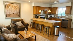 A modern kitchen with wooden cabinets, a central island with stools, a dining area with a leather sofa, and a painting on the wall. - Applewood Charming Two Bedroom Big Fenced Yard (Fort Collins)