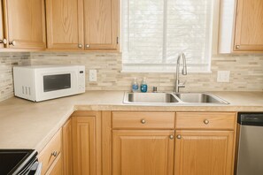 Una cocina con armarios de madera, un microondas blanco, un lavamanos de acero inoxidable y un lavavajillas.