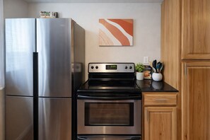 A kitchen with a stainless steel refrigerator, a black stove, wooden cabinets, and a wall-mounted abstract painting. - Quiet Cozy Downtown Cheyenne (Cheyenne)