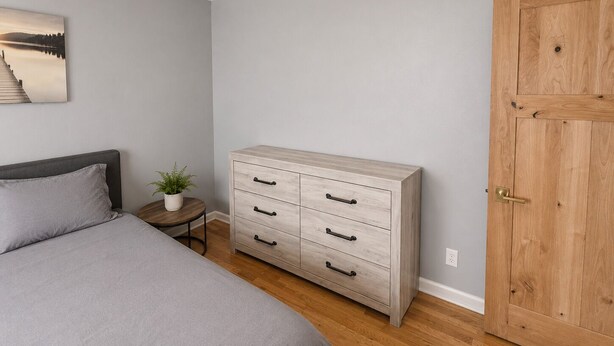 Une chambre à coucher avec un lit, une table de chevet ornée d’une plante, une commode en bois avec des tiroirs, une porte en bois et un ventilateur de plafond.