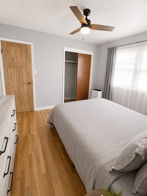 A bedroom with a bed, wooden floor, ceiling fan, closet, and window with curtains.