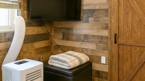 Quarto com televisão de tela plana fixada em parede de madeira, ar condicionado portátil branco e um banquinho preto com almofada.