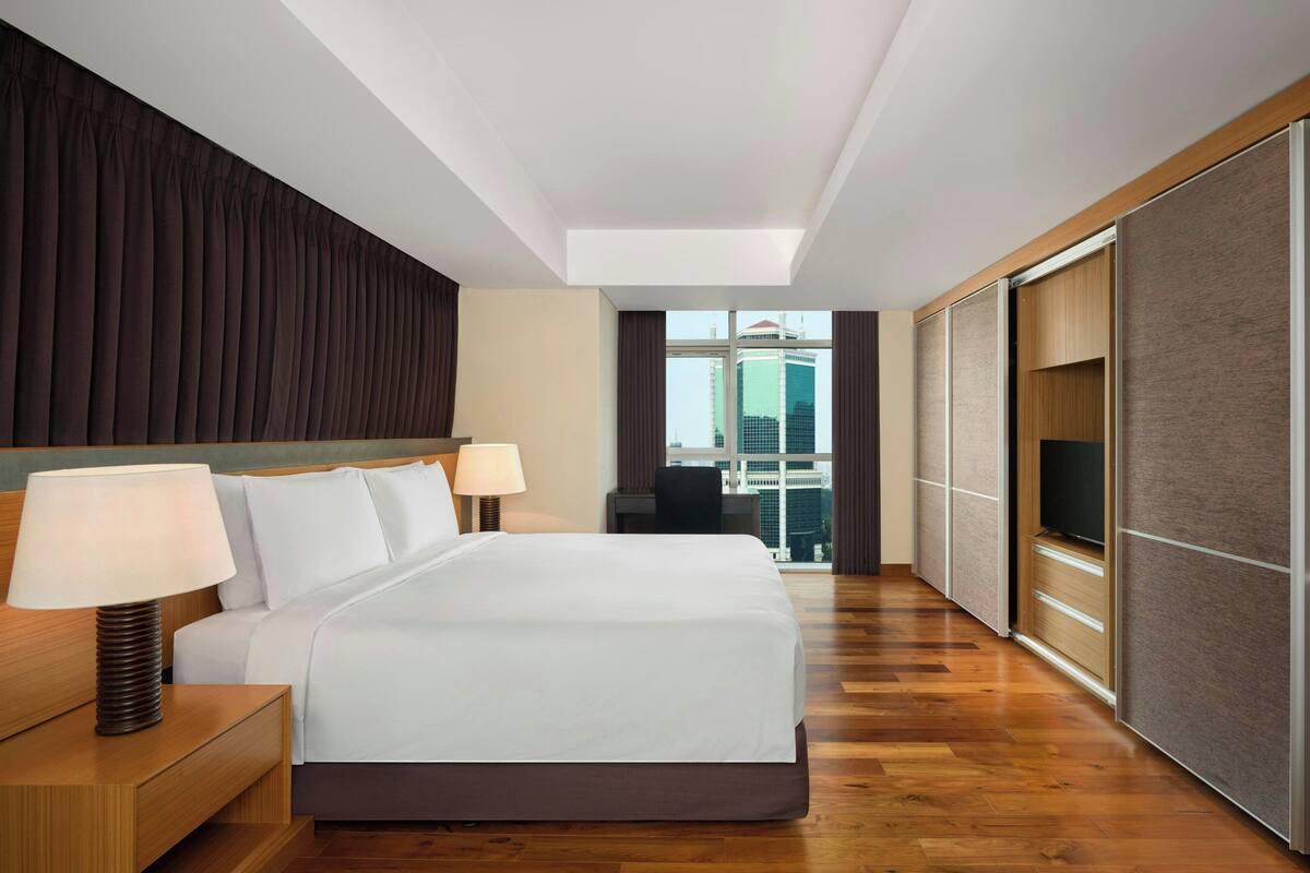 A modern hotel room with a large bed, bedside lamps, a TV, and a view of the city through a large window.