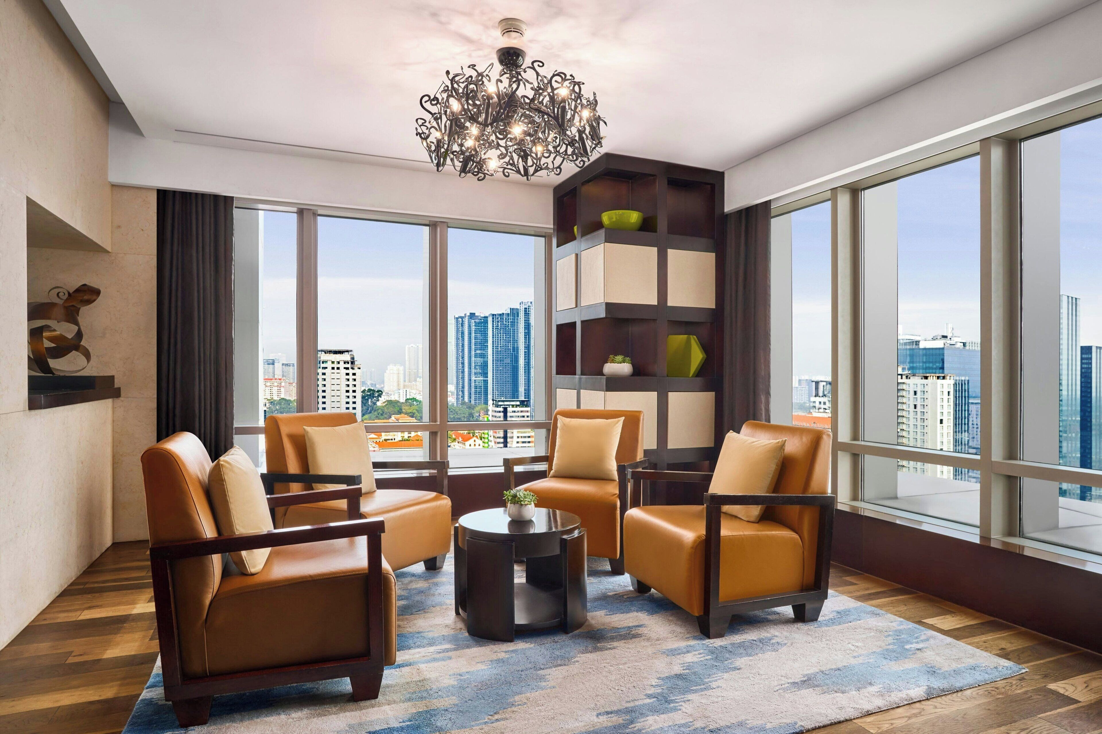 A modern living room with leather sofas, a round coffee table, and a decorative chandelier. Large windows offer a city view.