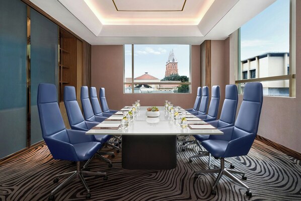 Un salón de conferencias con una larga mesa, sillas azules y vistas a un edificio con una torre.