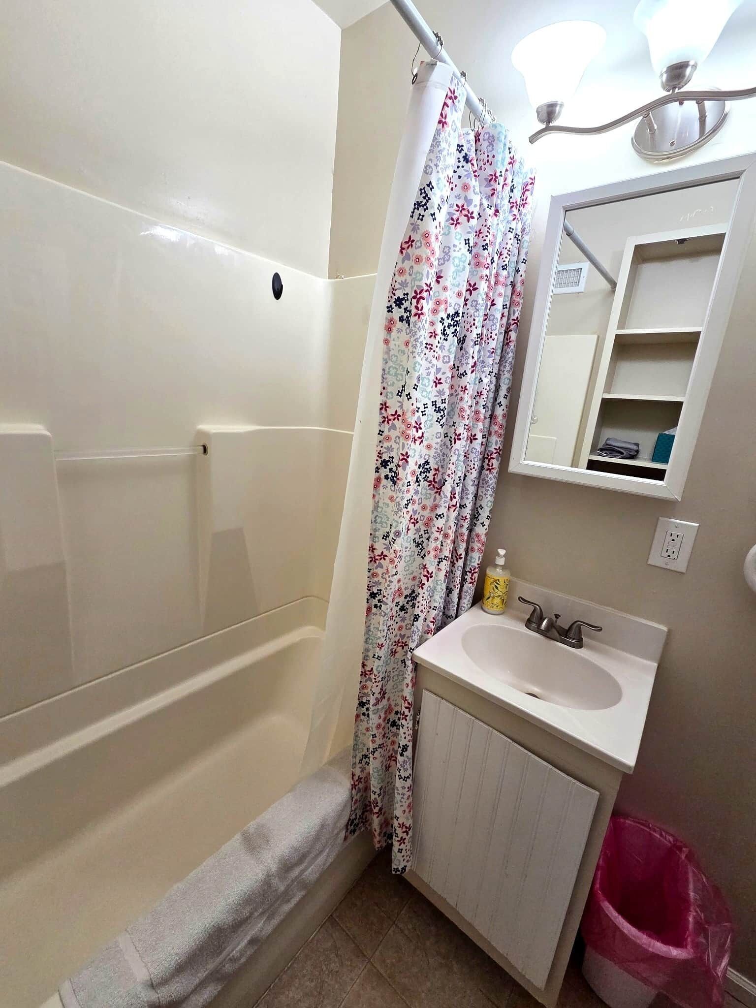 A bathroom with a bathtub, a shower curtain, a sink with a mirror above it, and a trash bin.