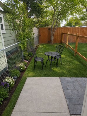 Un jardin avec une zone délimitée par une clôture, une clôture en bois, une table en métal avec des chaises et une terrasse en béton.