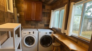 A compact laundry room with two washing machines, a wooden bench, and a window with curtains. - Cozy cottage in Cheyenne (Cheyenne)