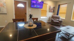 A modern living room with a glass-top dining table, a comfortable sofa, a television mounted on the wall, and a door with a decorative glass panel. - Cheyenne Frontier Homes #3 (Cheyenne)