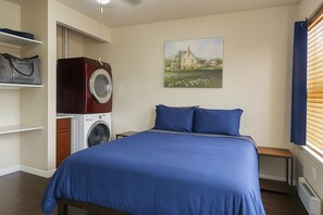 Un dormitorio con cama, lavadora, un cuadro y ventana con persianas.