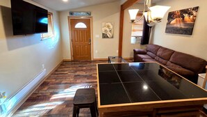 A living room with a wooden floor, a brown leather sofa, a glass-top coffee table, a TV mounted on the wall, and a door with a 'HOME' sign above it. - Cheyenne Frontier Apartment (Cheyenne)