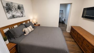 Une chambre à coucher comprenant un lit, des tables de chevet, une commode, un téléviseur et un couloir menant à une salle de bain.