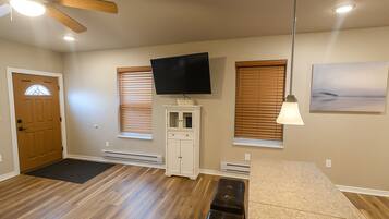 A spacious room with a wooden floor, a ceiling fan, a flat-screen TV mounted above a cabinet, and a large window with blinds.