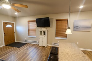 Una habitación amplia con piso de madera, ventilador de techo, televisor de pantalla plana sobre un mueble bajo y un ventanal grande con persianas.