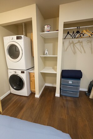 Une petite buanderie comprenant une machine à laver et un sèche-linge superposés, une étagère avec un bidon de lessive et un portant à vêtements avec des cintres.