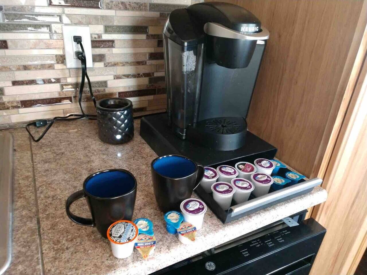 A kitchen with a coffee maker, mugs, and a container of yogurt cups.