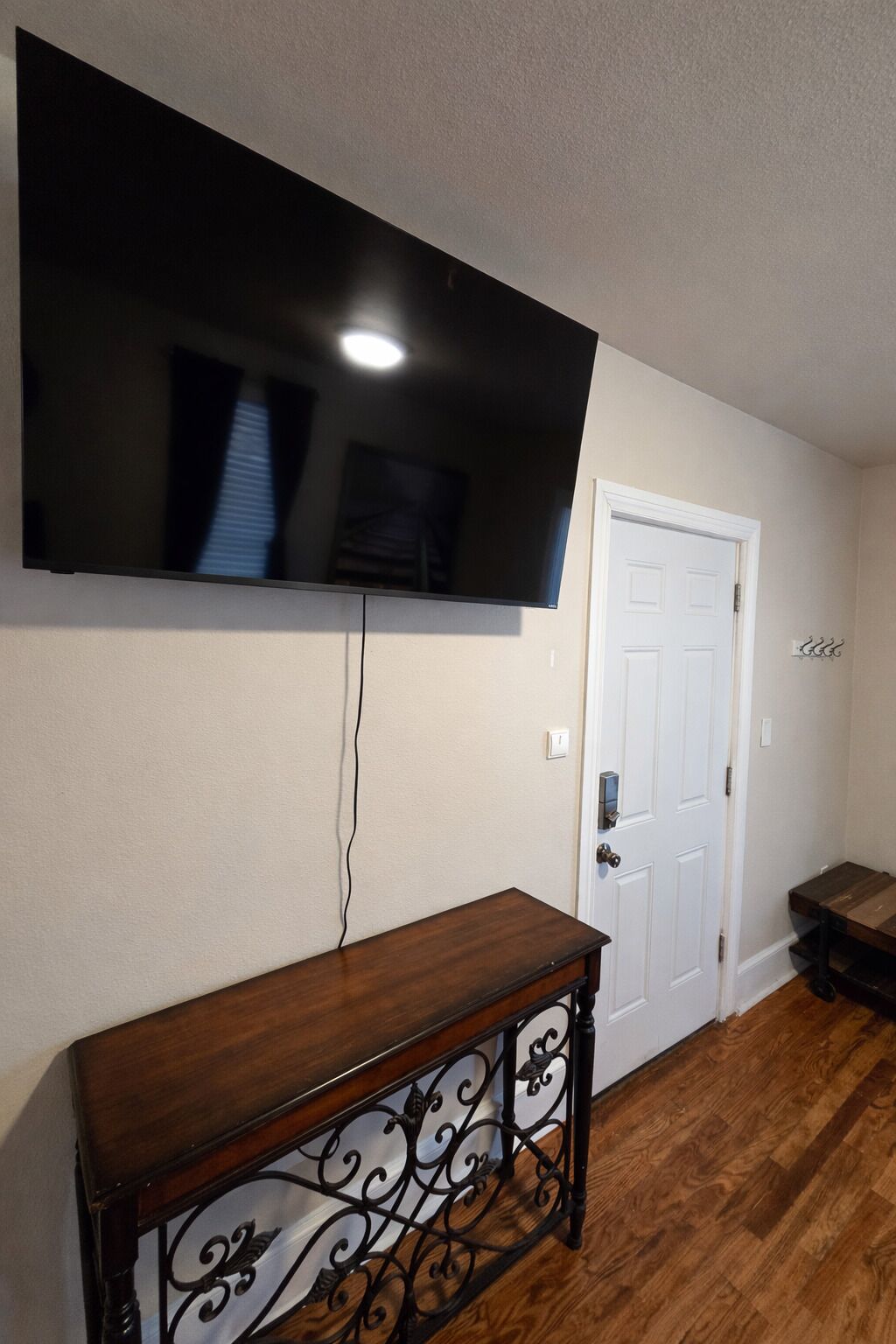 Een kamer met een wandgemonteerde flatscreen televisie, een houten console tafel met een decoratief metalen frame, en een witte deur met een deurklink.
