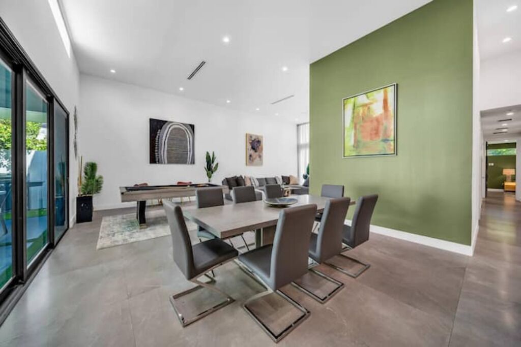 A modern dining area with a large table and chairs, a green accent wall, and artwork.