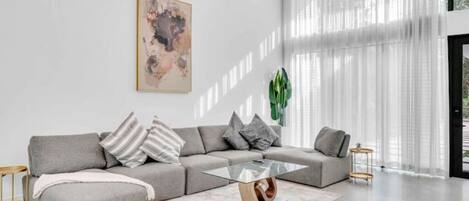 Una sala moderna con un sofá seccional gris, una mesa de centro de vidrio y un ventanal con cortinas traslúcidas.