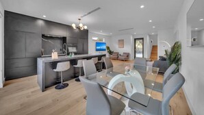 Una cocina moderna con una isla central, un comedor con mesa de vidrio y una sala de estar con sofá y televisor.