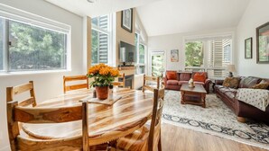 A spacious living room with a wooden dining table, a brown leather sofa, and a large window with a view of trees. - Bright & Cozy Getaway (Park City)