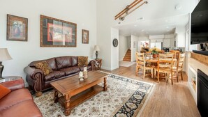 A spacious living room with a brown leather sofa, wooden coffee table, and a dining area with wooden chairs and a table. There are framed pictures on the wall, a staircase leading to another level, and a kitchen area visible in the background. - Bright & Cozy Getaway (Park City)