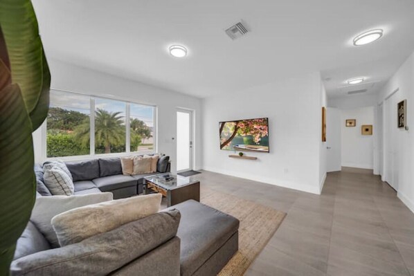 Un salon moderne avec un canapé gris, une table basse et un téléviseur fixé au mur. Une grande fenêtre offre une vue sur la verdure à l’extérieur.