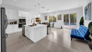 A modern kitchen with white cabinetry, a central island, and a dining area with a blue sofa. - Waterfront Estate•7BR•Heated pool•Spa•BBQ•Basketball (Miami)