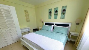 A bedroom with a bed, a nightstand, a chair, a wardrobe, and three wall art pieces. - Garden View Apartments - Managed by David & David Ltd (Roseau)