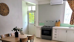 A kitchen with a dining table, chairs, a clock, an air conditioner, and a window with curtains. - Garden View Apartments - Managed by David & David Ltd (Roseau)