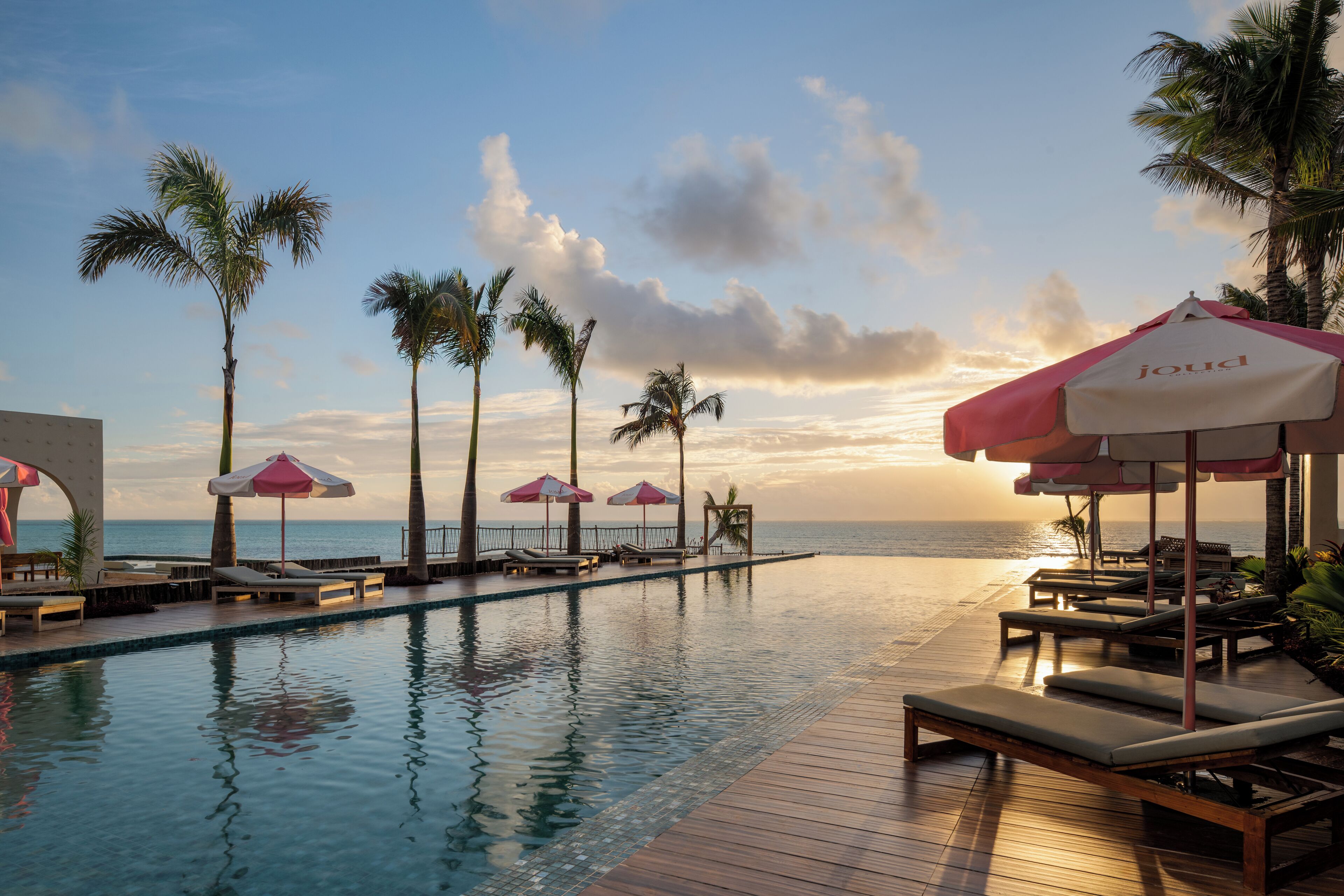 An infinity pool with lounge chairs and umbrellas, palm trees, and a view of the ocean.
