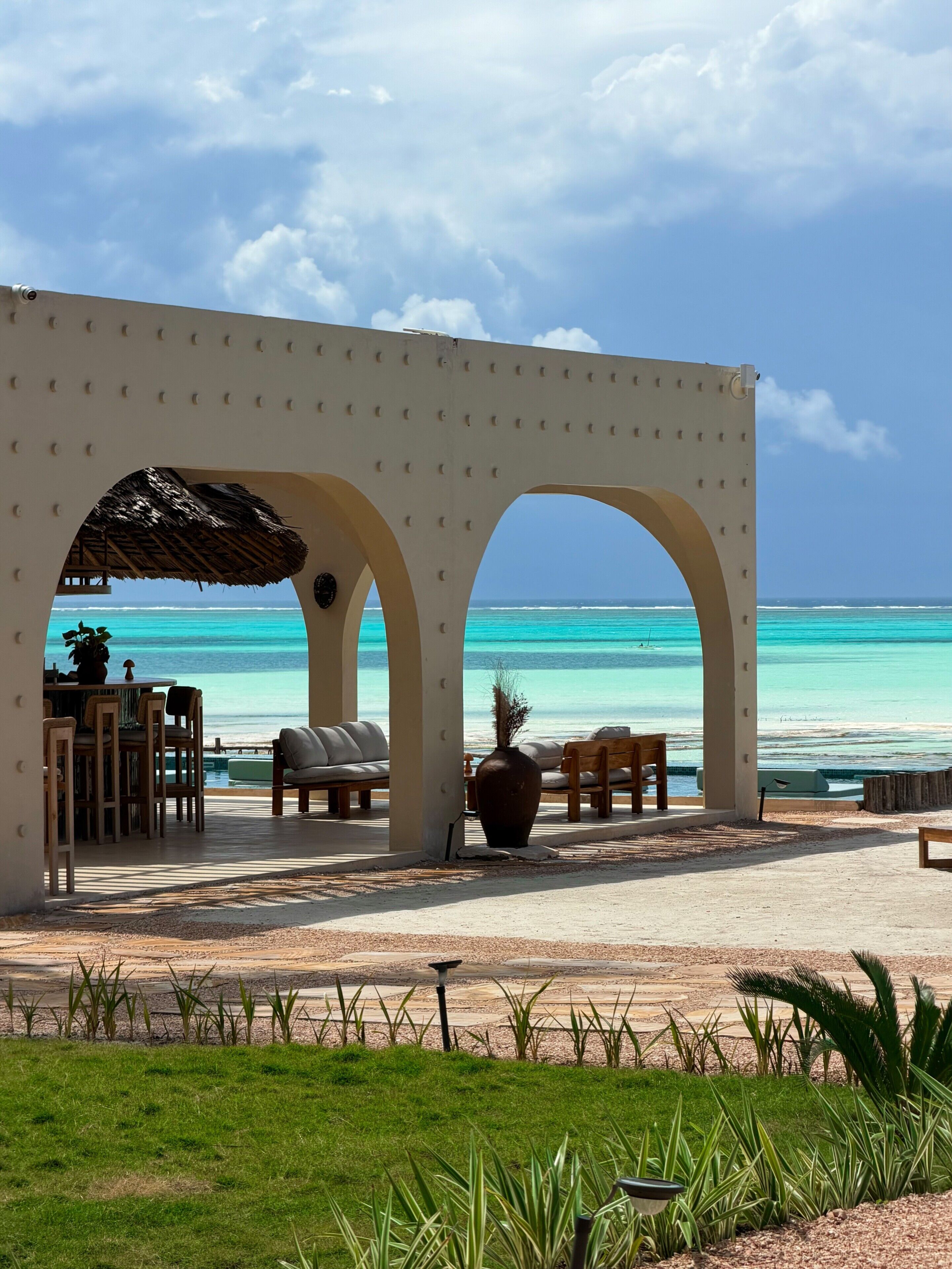 A beachside lounge with a view of the ocean, featuring a thatched roof, seating, and a large vase.
