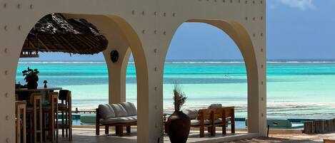 A beachside lounge with a view of the ocean, featuring a thatched roof, seating, and a large vase.